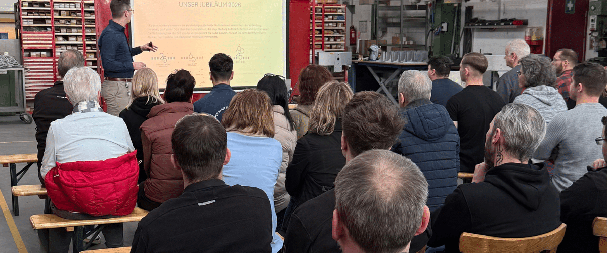 Bild von der Betriebsversamlung, Mitarbeiter*innen sitzen in Reihen hintereinander, vorne steht ein Beamer und eine Leinwand und der Chef der spricht.