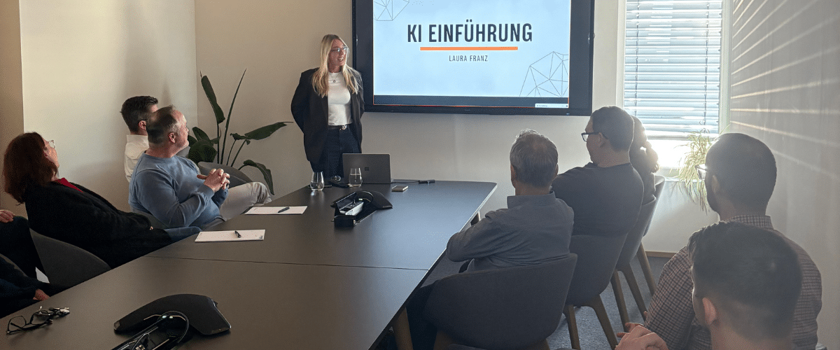 Eine Person hält eine Präsentation in einem Konferenzraum mit einem großen Bildschirm. Es sitzen einige Personen an einem langen Tisch und hören zu.