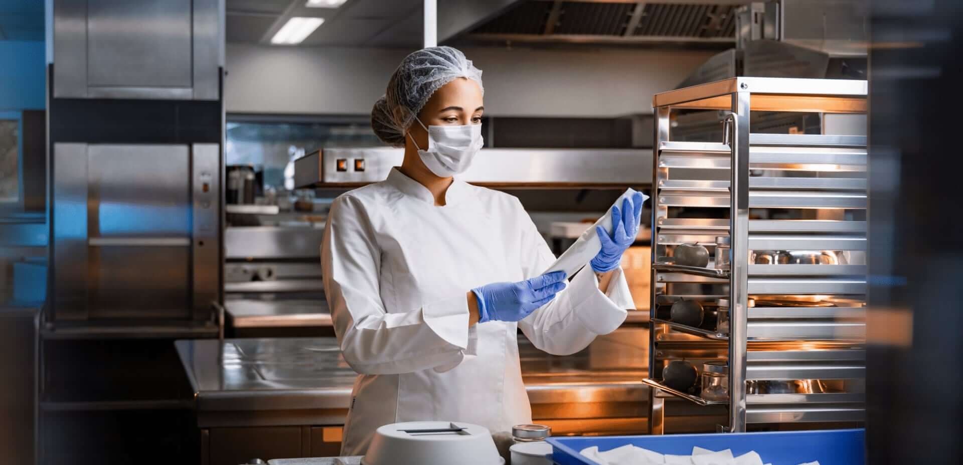 Damen mit Mundschutz und Handschuhen hält eine Serviettenpapierwicklung mit Besteck in der Hand. Im Hintergrund steht ein Tablettwagen.