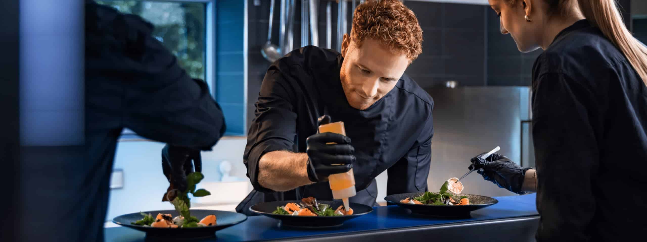 Zwei Damen und ein Herr am Anrichteband sind konzentriert und richten die Teller mit Essen an.