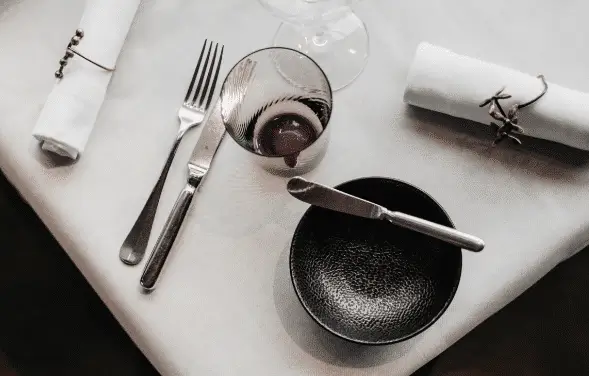 Eleganter gedeckter Tisch mit einer Gabel, einem Messer und einem Glas neben einer schwarzen Schüssel mit einem Buttermesser obenauf. Zwei weiße Servietten mit dekorativen Ringen liegen auf einem weißen Tischtuch.