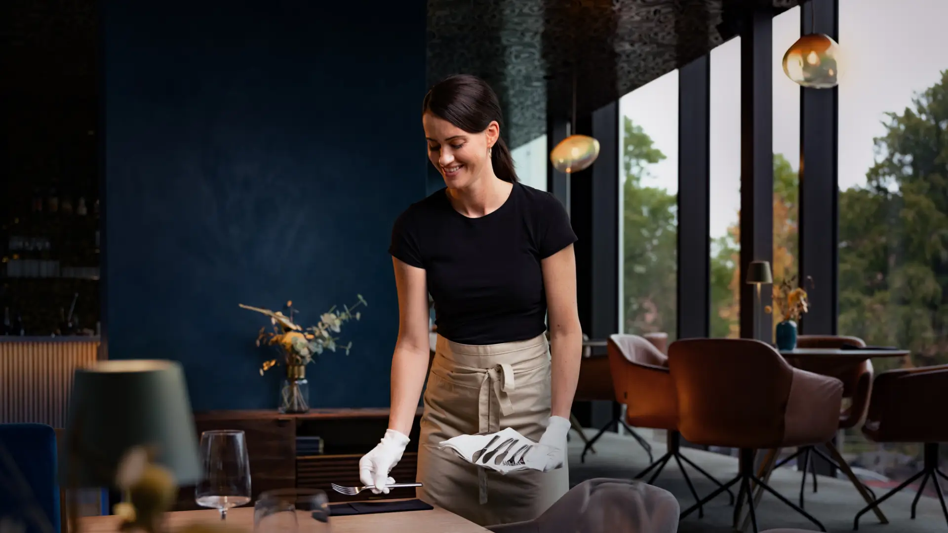 Eine Dame mit einer Schürze steht an einem Tisch und deckt diesen mit Besteckteilen ein. Im Hintergrund sind weitere Tische des Restaurants.