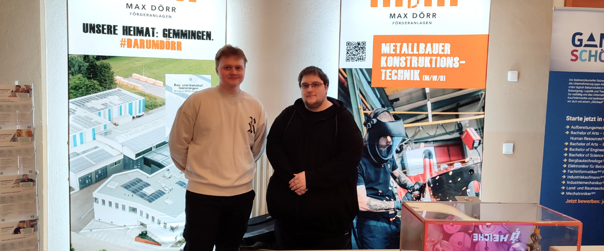 Zwei Personen stehen an einem Stand auf einer Ausbildungsmesse. Im Hintergrund zwei beleuchtete Rollups mit Aufdruck, vorne einen Tisch mit GiveAways.