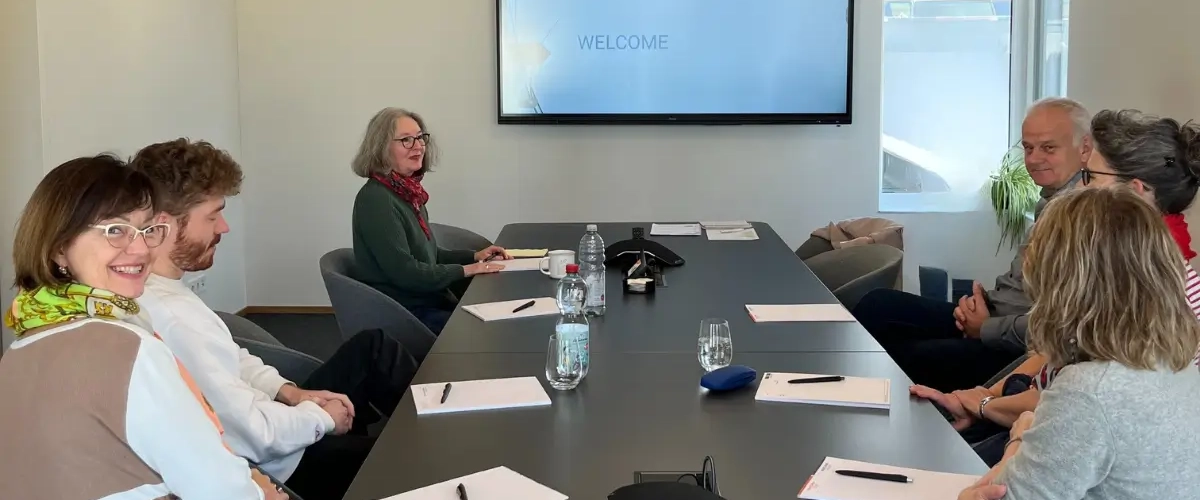 Eine Gruppe von Personen sitzt im besprechungsraum an einem langen TIsch und auf dem Bildschirm ist einen Titelfolie mit den Worten "Welcome"