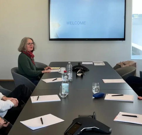 Eine Gruppe von Personen sitzt im besprechungsraum an einem langen TIsch und auf dem Bildschirm ist einen Titelfolie mit den Worten "Welcome"