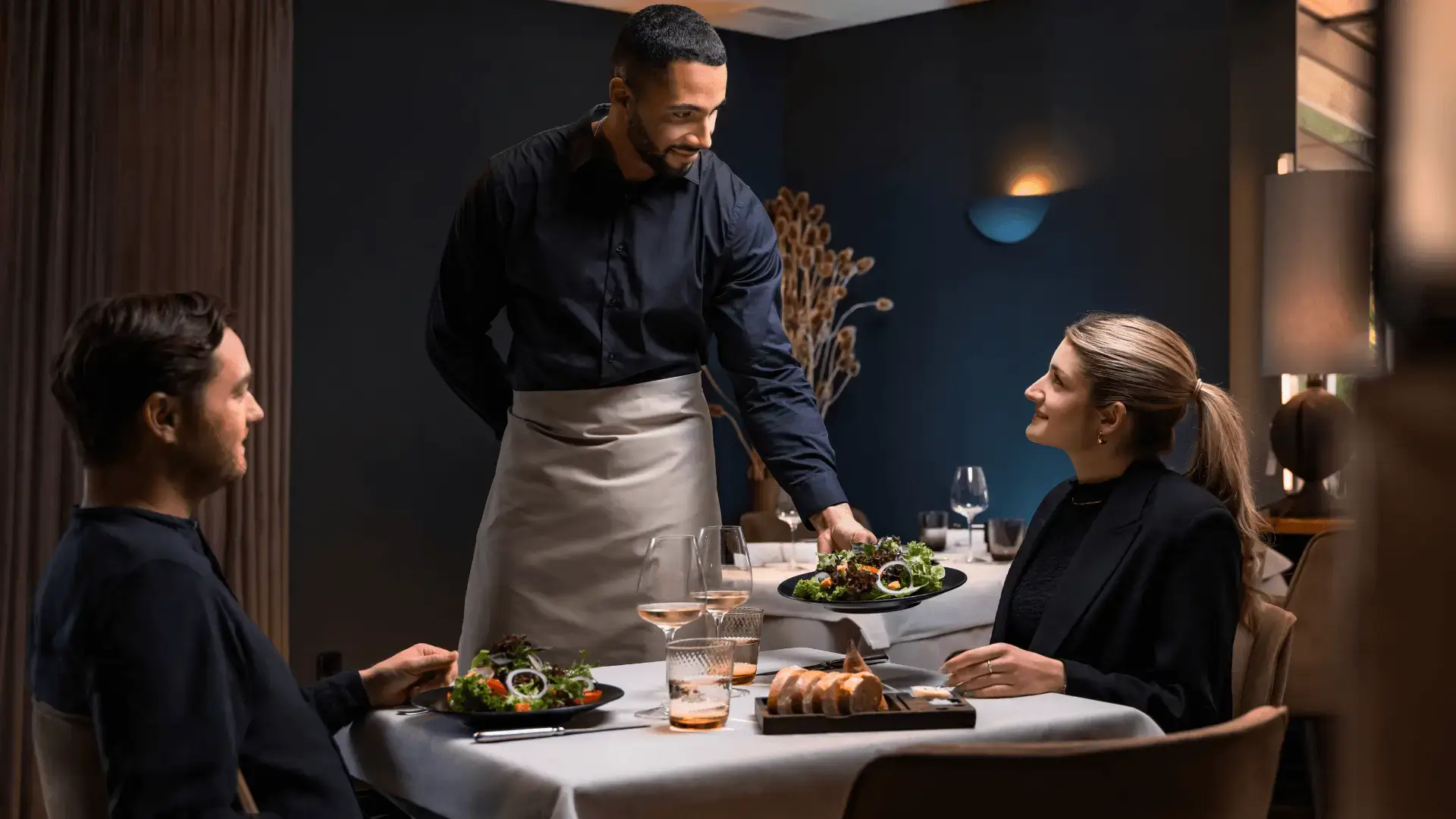 Zwei Personen (männlich, weiblich) sitzen in einem Restaurant an einem Tisch. Vor ihnen steht ein Glas Wein und jeweils ein Salat und etwas Brot. Der Kellner steht neben dem Tisch und reicht der Dame gerade ihren Salatteller.