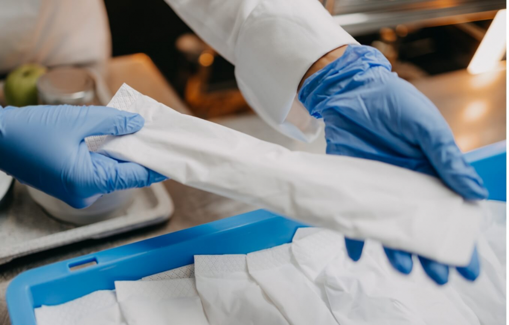 Zwei Personen mit blauen Handschuhen hantieren mit einem eingewickelten Gegenstand, möglicherweise Utensilien, über einem blauen Tablett, das mit ähnlichen eingewickelten Gegenständen gefüllt ist, in einer Einrichtung, bei der es sich anscheinend um einen Lebensmittelladen oder eine medizinische Einrichtung handelt.