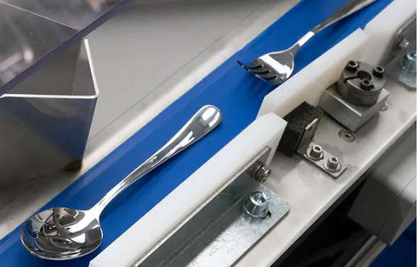 Nahaufnahme einer automatisierten Maschine mit einem Metalllöffel und einer Gabel, die nebeneinander auf einem blau-weißen Fördermechanismus liegen, mit verschiedenen mechanischen Komponenten in der Nähe.