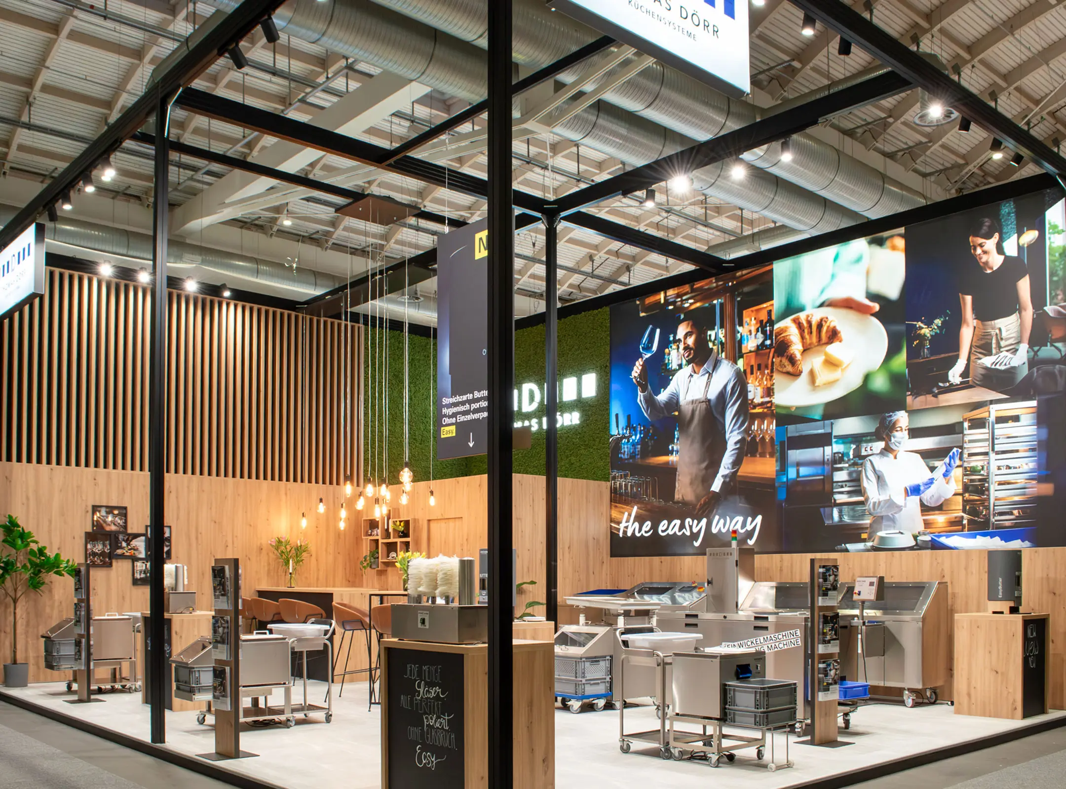 Ein moderner Messestand mit hölzernen Akzenten, Hängelampen, Küchengeräten und großen Wanddisplays, die Köche und Szenen der Essenszubereitung bei heller Innenbeleuchtung zeigen.