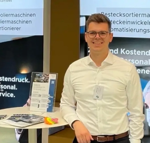 Person steht an einem kleinen Messestand an einem Stehtisch mit Giveaways und schaut in die Kamera.