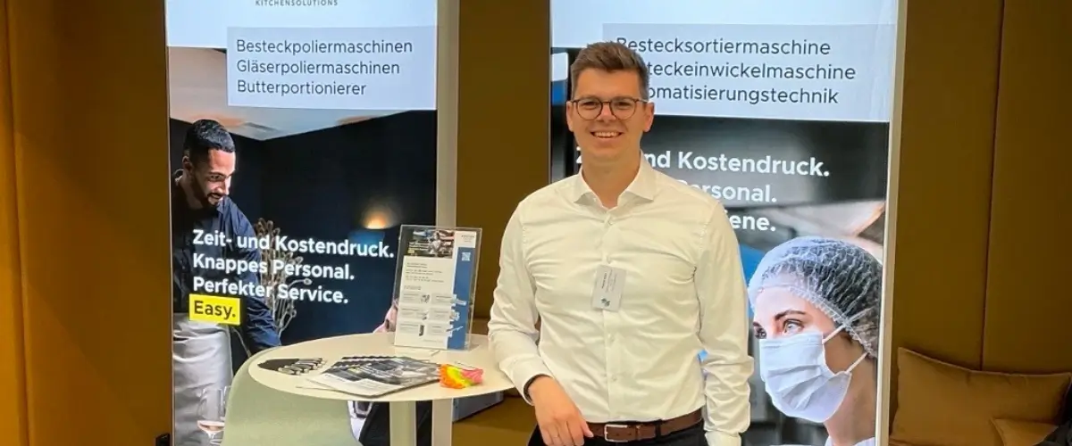 Person steht an einem kleinen Messestand an einem Stehtisch mit Giveaways und schaut in die Kamera.