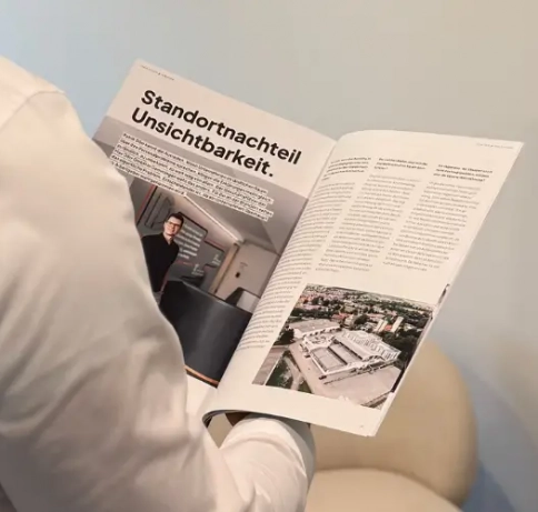 Person von hinten hält ein aufgeschlagenes Magazin in der Hand, rechts im Bild ist noch eine Pflanze zu sehen. 