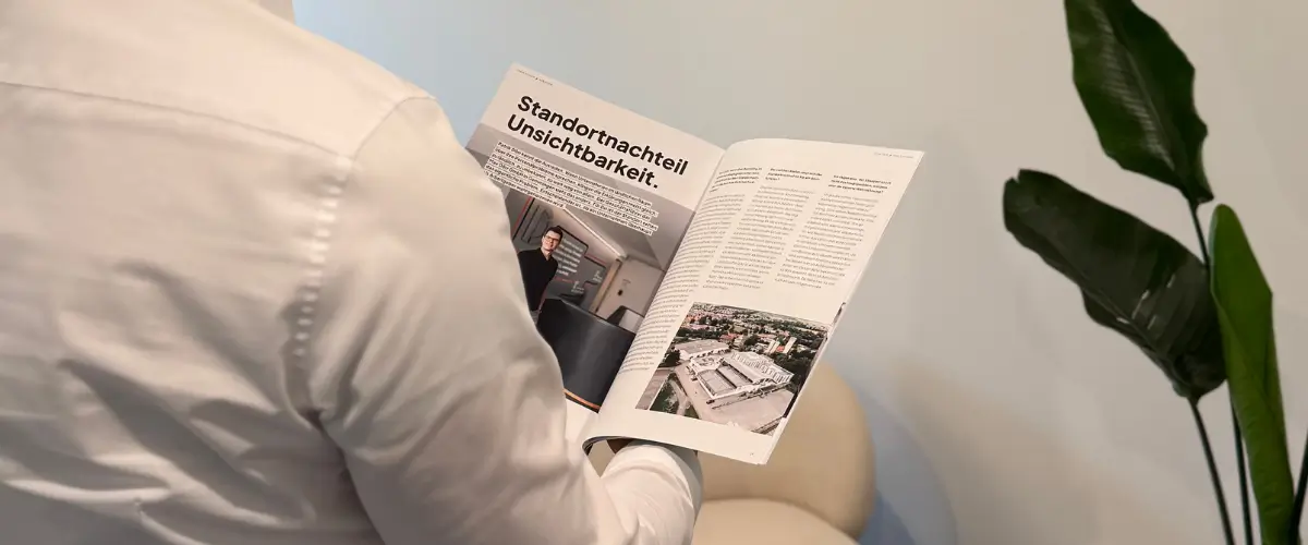 Person von hinten hält ein aufgeschlagenes Magazin in der Hand, rechts im Bild ist noch eine Pflanze zu sehen.