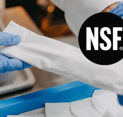 Zwei Hände mit Einweghandschuhen halten eine Serviettenwicklung unserer Besteckeinwickelmaschine, die Besteck einwickelt. Darüber ist ein NSF-Logo platziert.